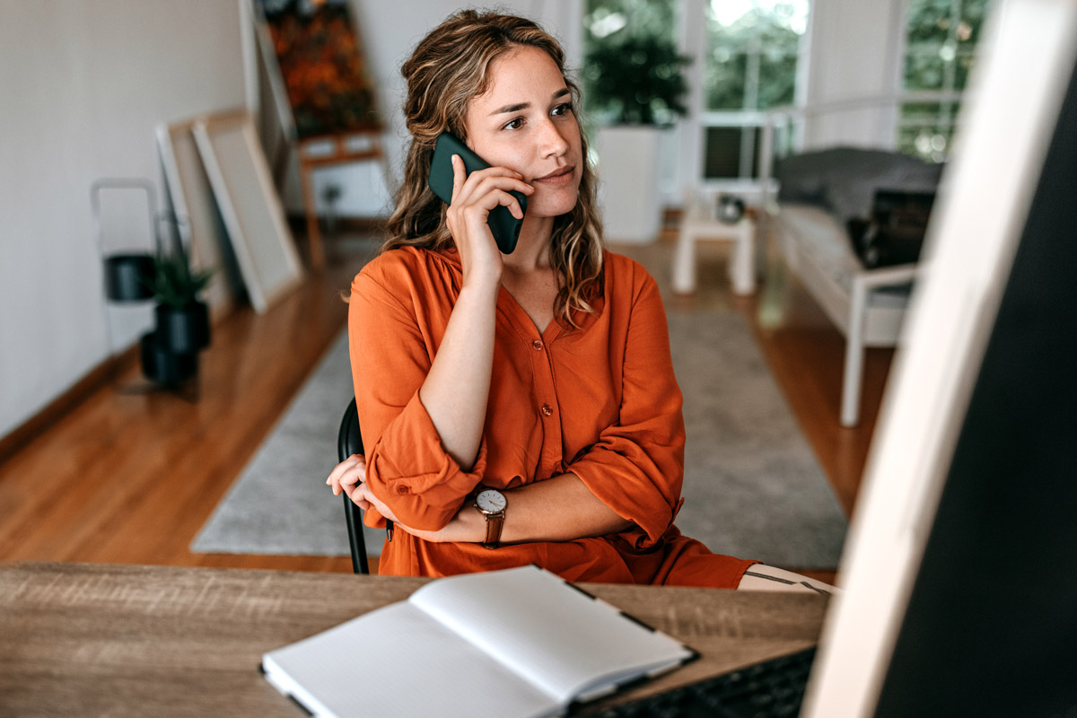 A woman makes a phone call while sitting at her desk at home.