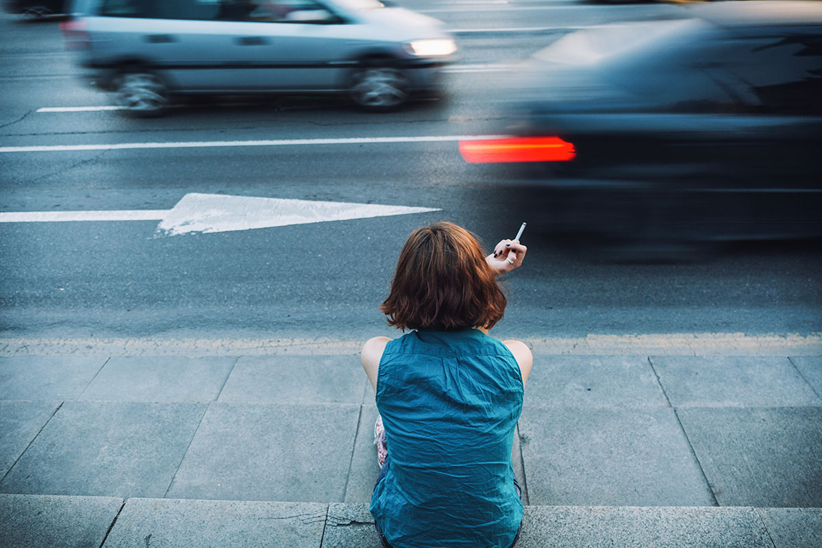 girl smoking on a sidewalk near the boulevard. Long exposure image with blurred car and blue dark toning.