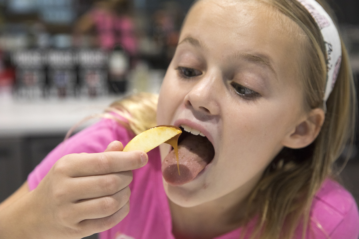 A girl with blonde hair and blue eyes eats a sliced apple dipped into caramel sauce.