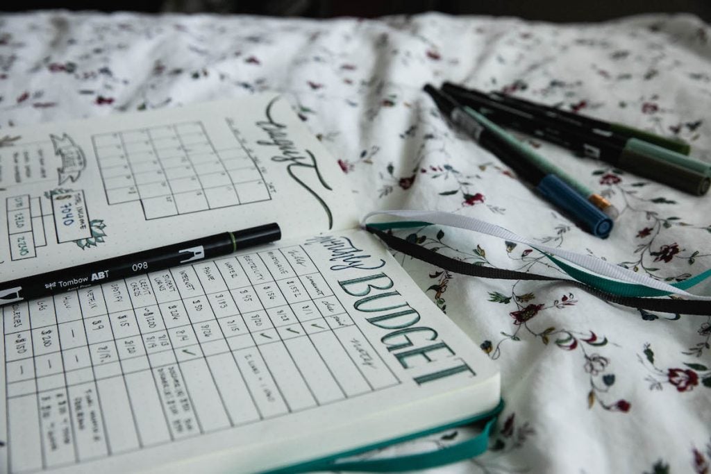 An open budget journal is shown lying on top of floral sheets, alongside several pens.