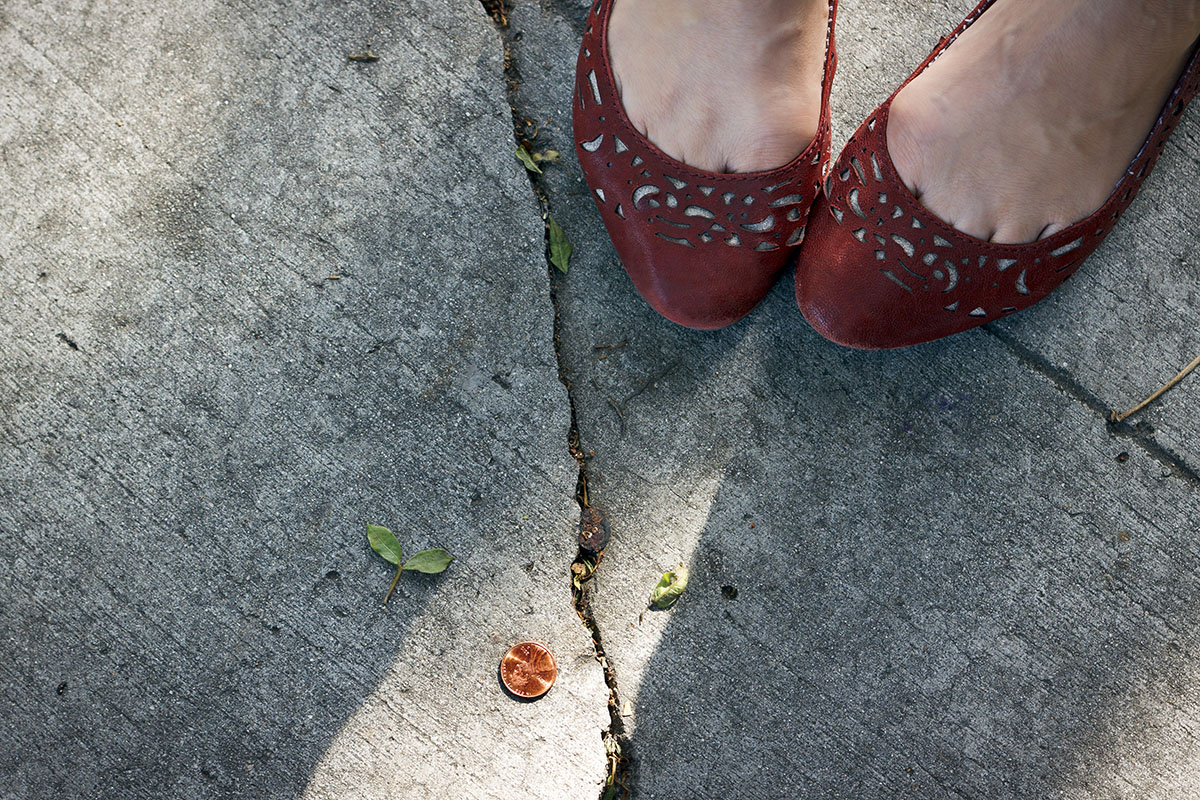 A girl in red shoes finds a penny on the sidewalk.