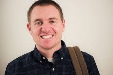 A portrait of a young, white mail wearing a flannel shirt and a book bag over his shoulder.
