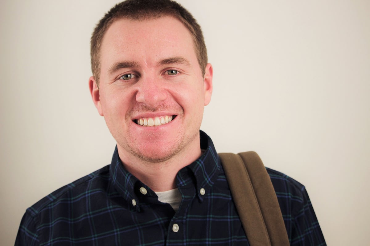 A portrait of a young, white mail wearing a flannel shirt and a book bag over his shoulder.