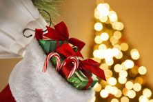 A Christmas stocking filled with goodies hangs from a fireplace mantelpiece while the lights from Christmas tree glow out of focus in the background.