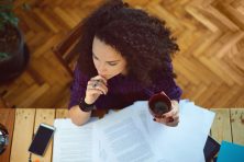 Young woman working at home, holding cup of coffee, and reading something.