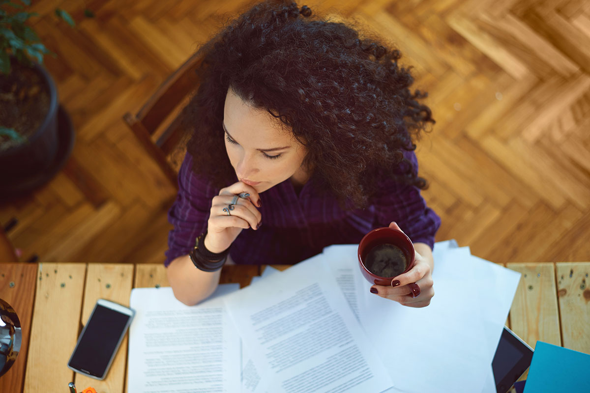 Young woman working at home, holding cup of coffee, and reading something.