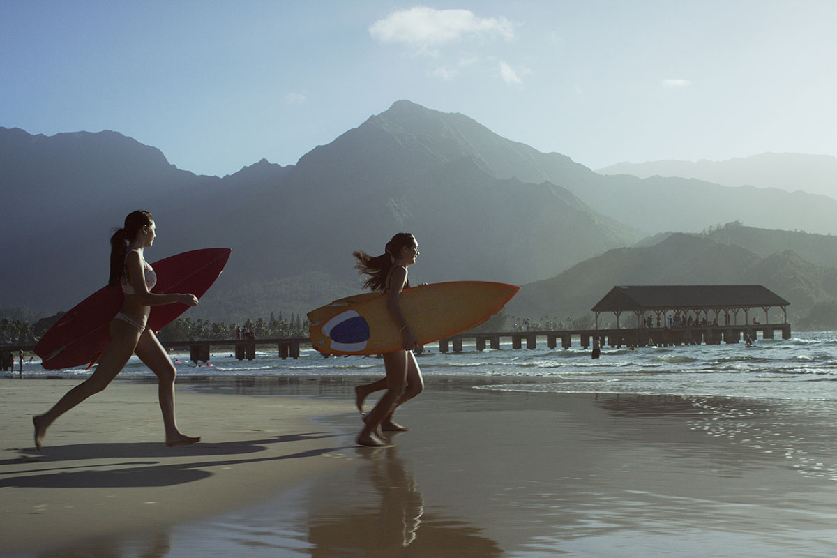 Three people on a beach, running into the ocean with surfboards in a tropical climate, with mountains in the background.
