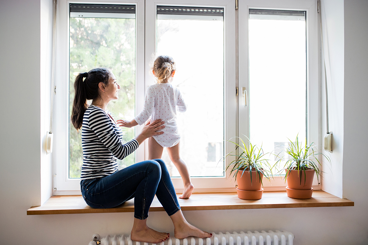 A woman helps her daughter stand as they look out the window.