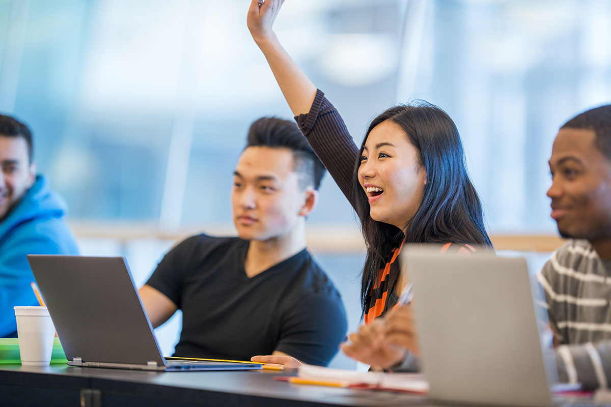 college students in class while one girl has hand raised to answer a question