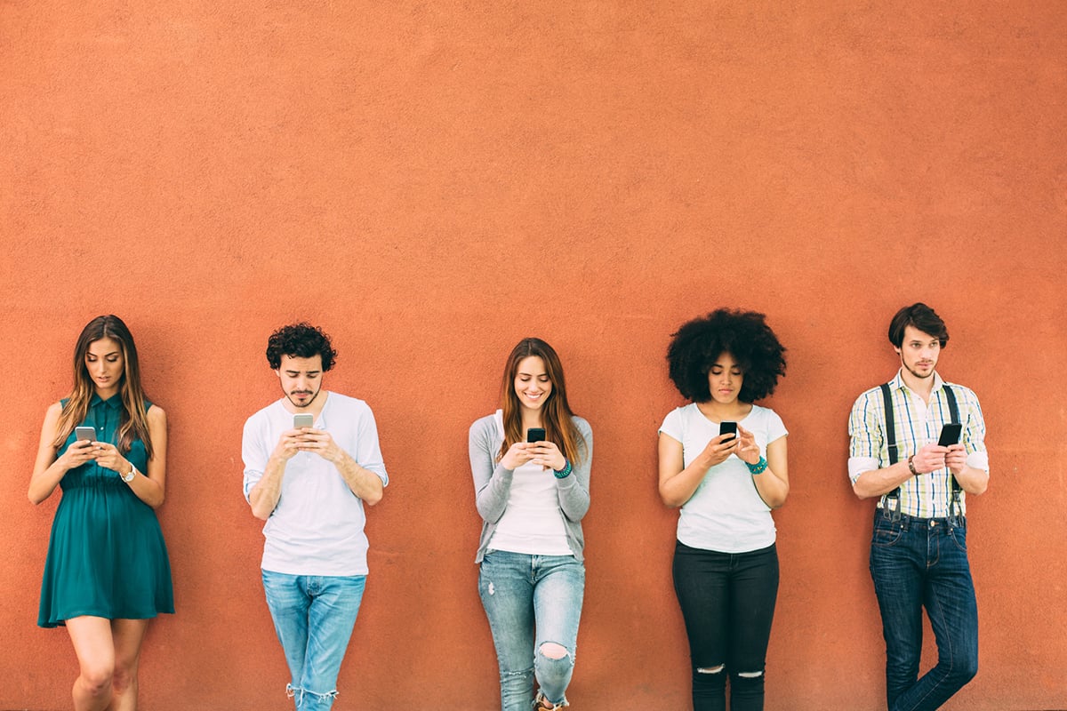 Five people texting holding cell phones while leaning against an orange wall