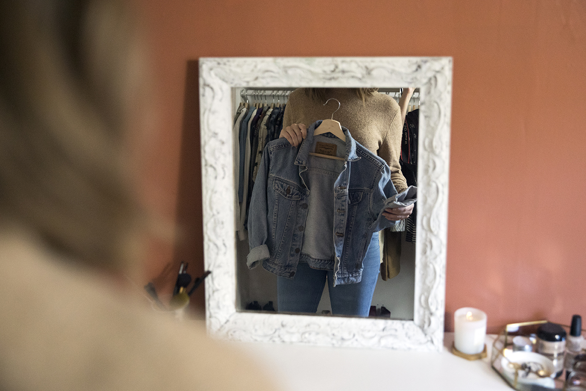 A woman looks at a jean jacket through a mirror in her closet.