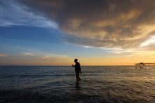 A woman walks through in the ocean as the sun sets.