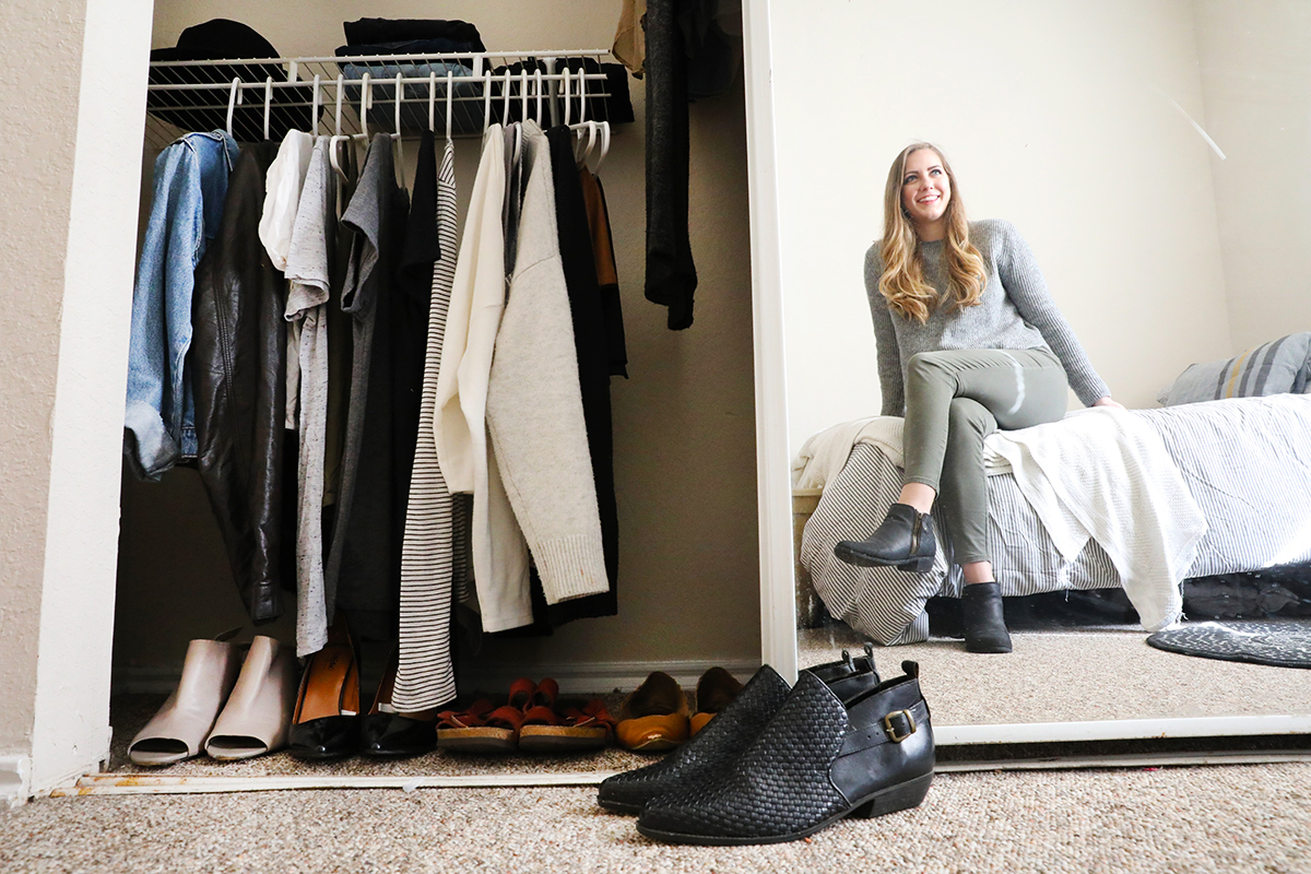 A woman is reflected in a wall mirror sitting on the bed with her closet in the foreground