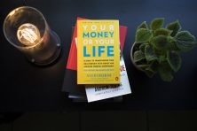 A stack of financial books sit on a black desk surrounded by a plant and lamp.