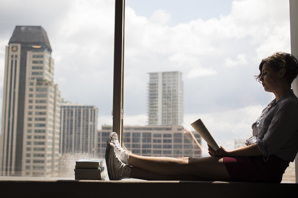 A woman reads a book while sitting on a window.