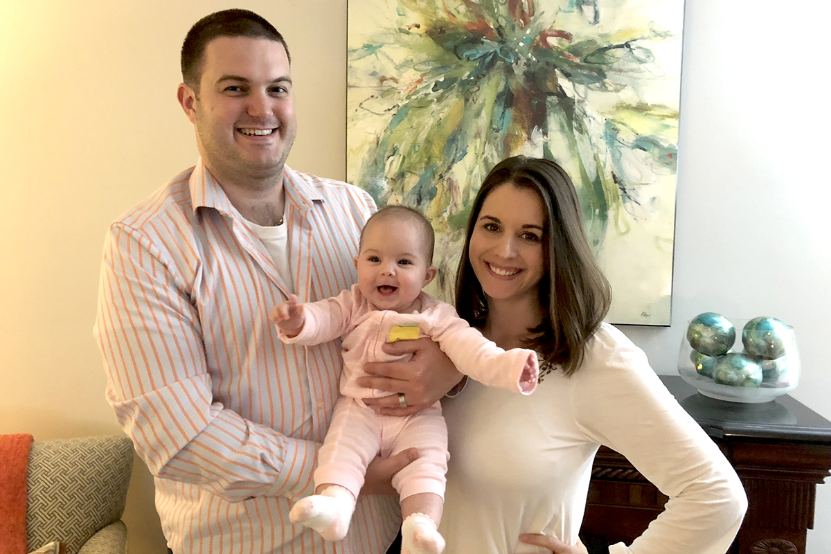 A husband and wife stand next to each other as they pose for a picture. The husband is holds up their baby.