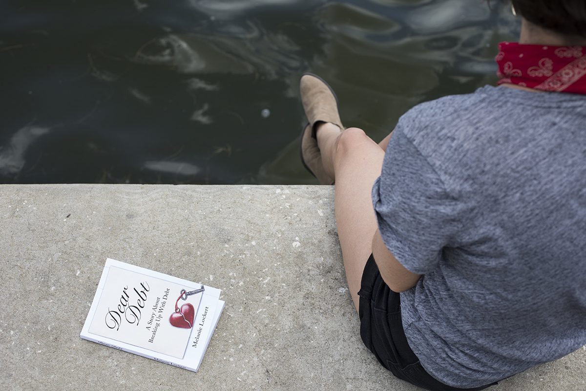 A woman sits on a sea wall while reading a book about debt.