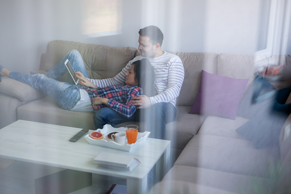 Young Couple Relaxing On Sofa, Chatting Smiling, Changing Channels And Surfing Internet.