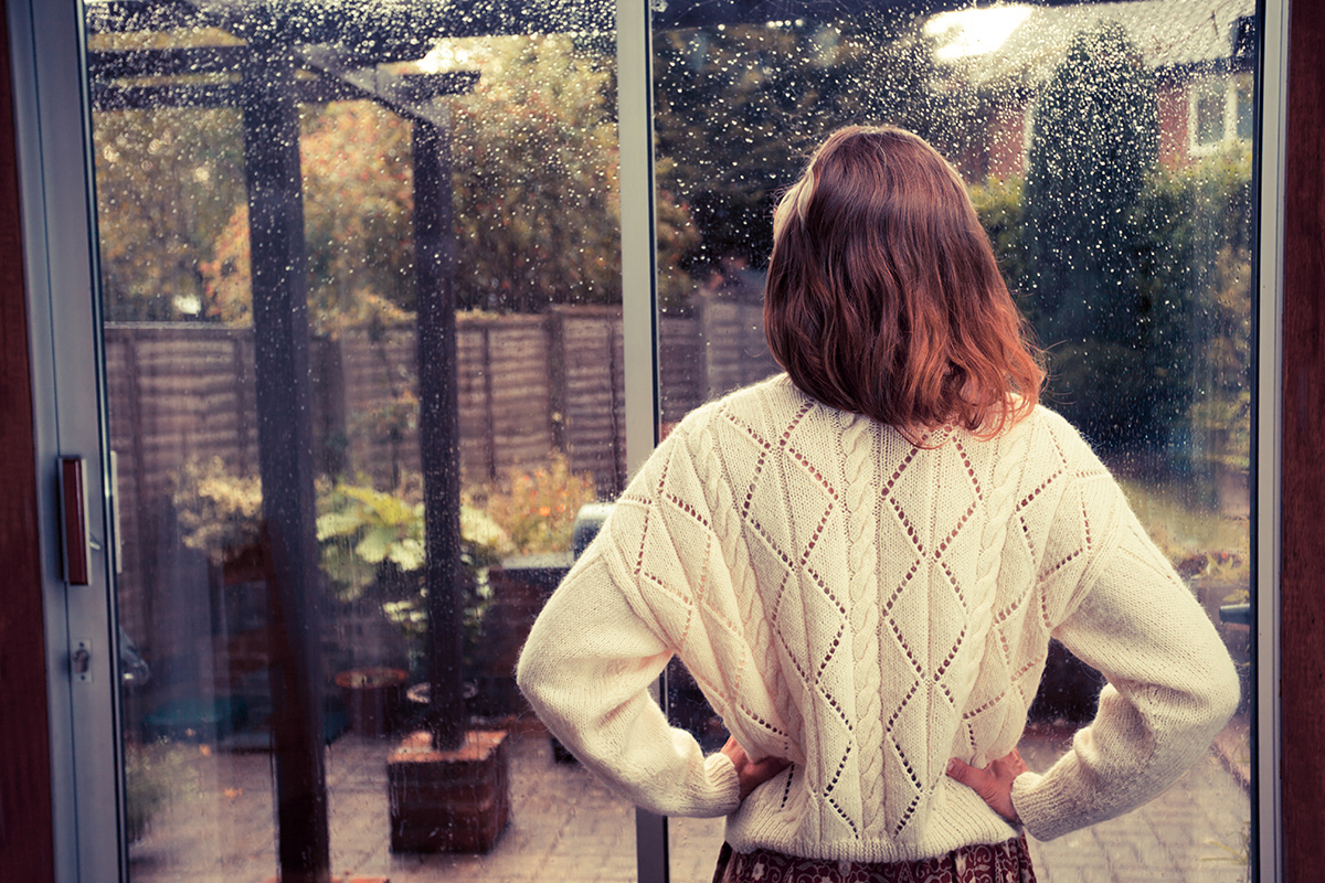 A young woman is standing by the french doors in her house and is looking out at the rain