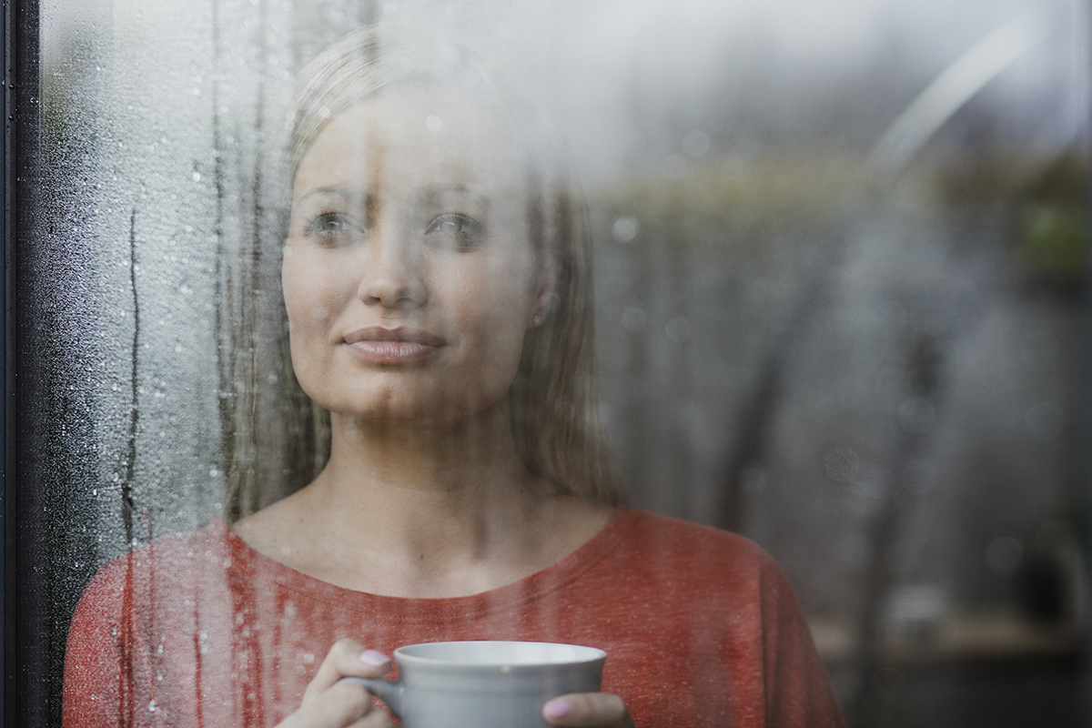 Woman is drinking a cup of tea while looking out of a window in her home.