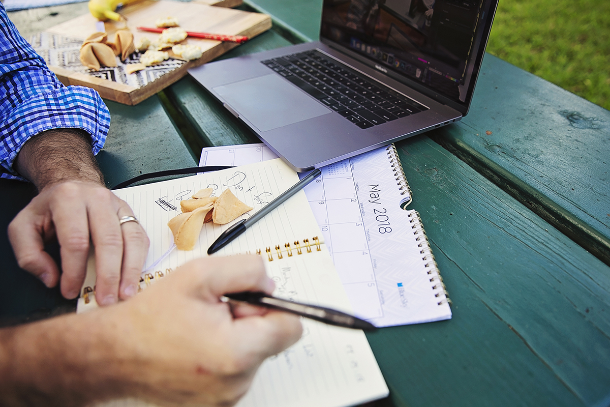 A man with a laptop writes in a notebook while eating at a picnic table.
