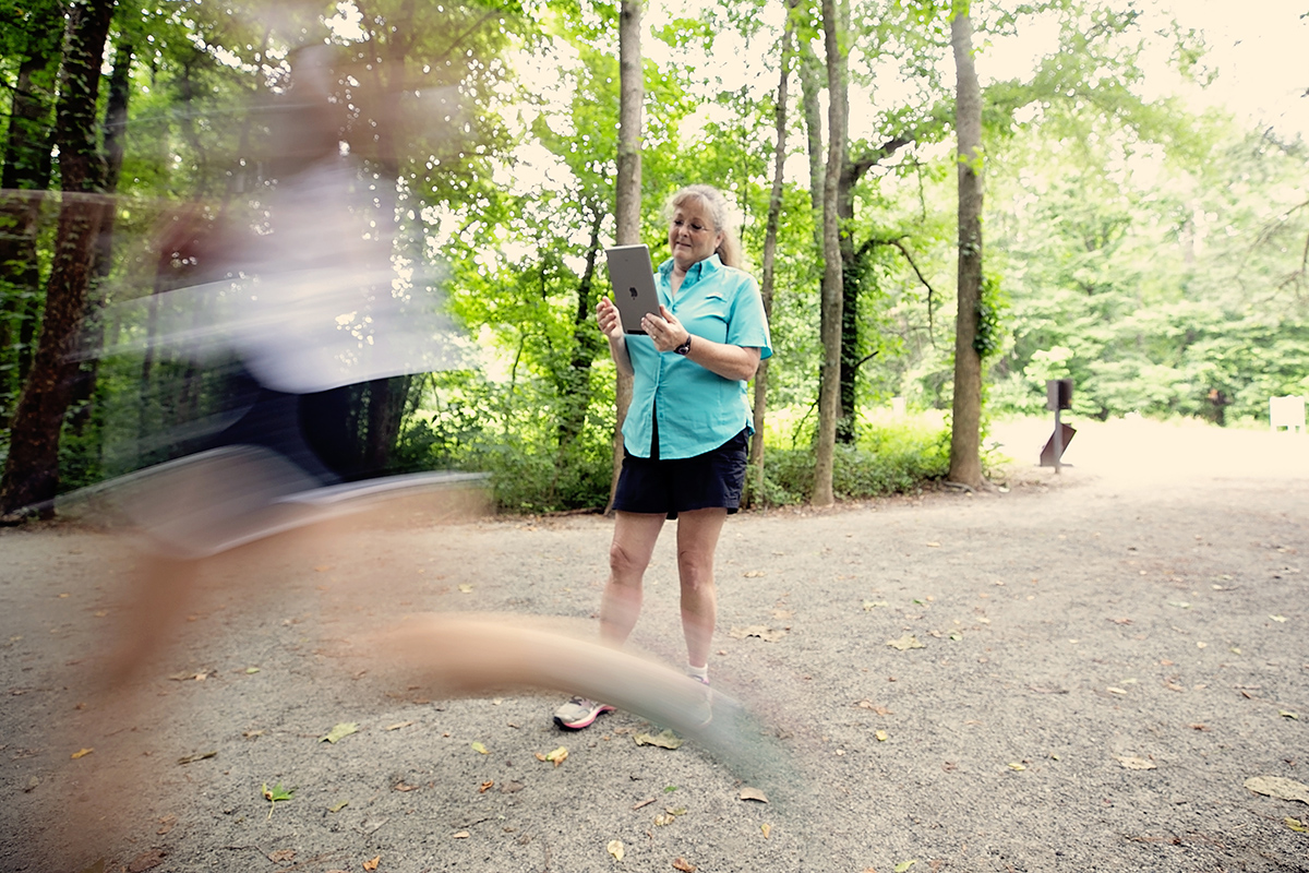 a woman runs past another woman, her fitness trainer, on a path in the woods.