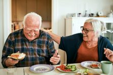 A retired couple laugh at the kitchen table in their home while eating breakfast.