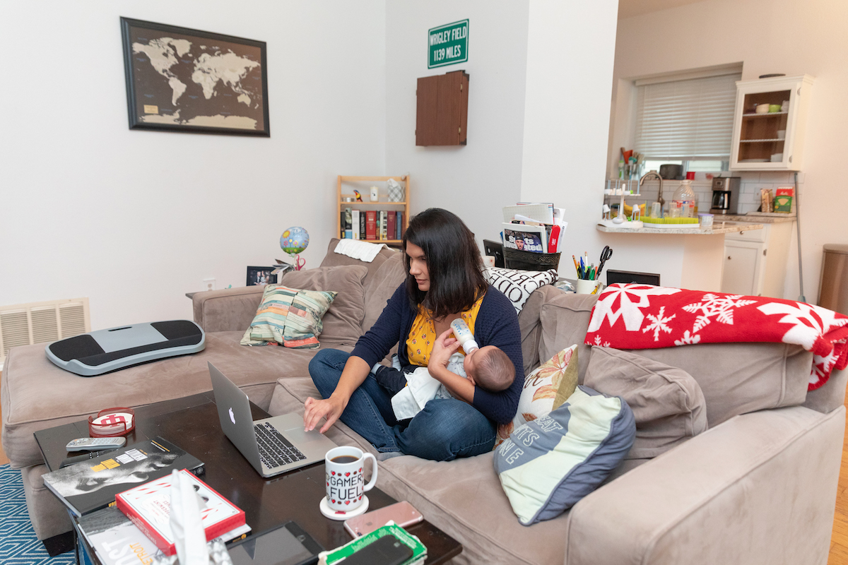 Laura Niebauer works on her laptop while holding her infant son at home.