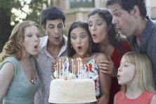 A woman surrounded by friends prepares to blow out birthday candles.