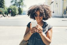 A young woman walks down the street. She wears sunglasses and is smiling down at her phone screen.