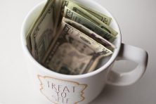 A coffee cup with a lot of money in it sits on a plain white surface.