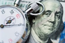 A stopwatch sits next the face of Ben Franklin on a $100 dollar bill.