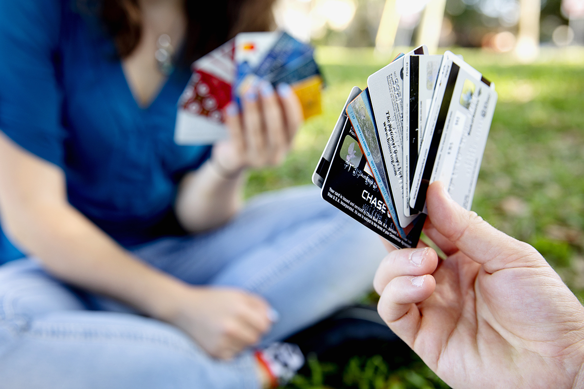 Two people sit in the grass, each holding a fan of credit cards as if they are playing a card game.