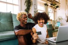 Two gay men look happy while sitting on the floor holding a laptop and credit card.