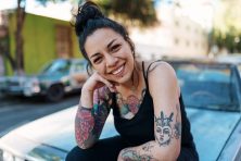 A woman with tattoos smiles as she sits on the hood of a car.