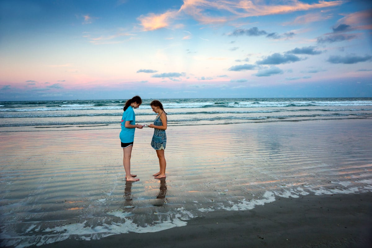 Two teenage girls look at their phone while standing in the water at Coca Beach during sunset. 