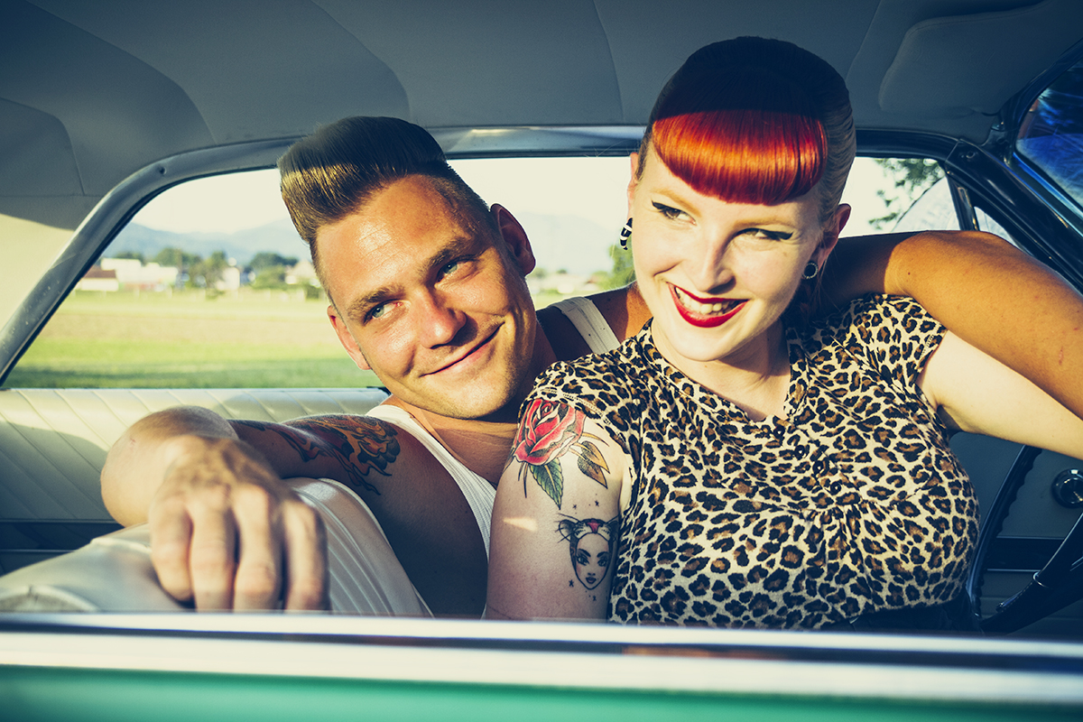 An interesting retro rockabilly couple smile while sitting in a car.