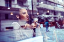 A woman seen through a window drinks coffee.
