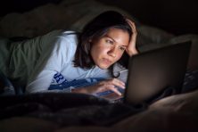 A young woman checks her computer while laying in bed.