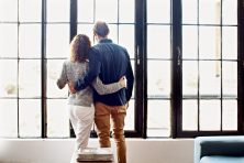 A married couple look out of windows in their home.
