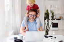 A boy sits on his dad shoulders, playing with his father's ears as his father works from home.