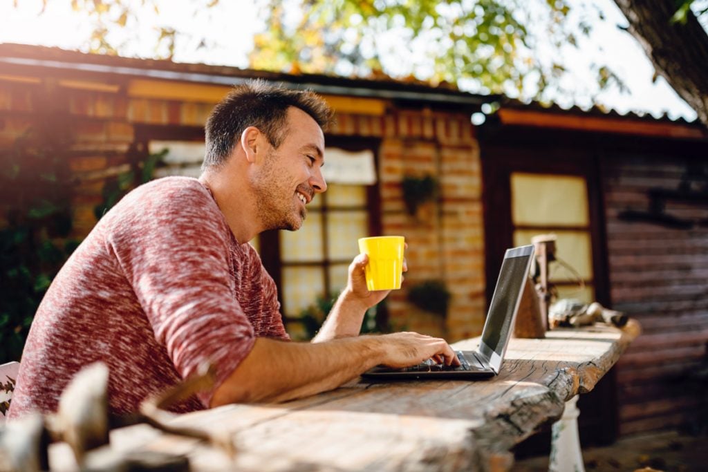 A man works on his laptop outside while sipping coffee from a bright yellow mug. The house behind him is made of brick.