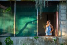 A woman stands for a portrait in the busted window of an ugly home that&rsquo;s inhabitable.