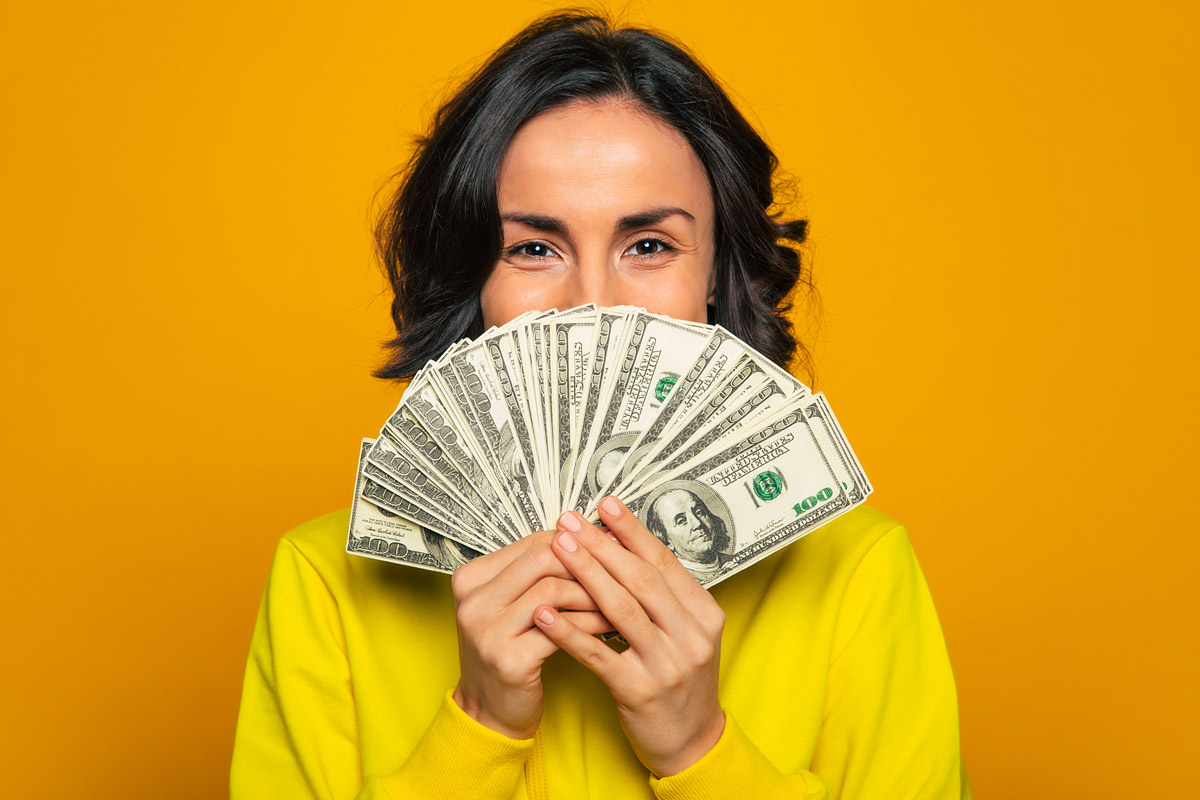 A woman smiles as she shows off her money. She's wearing yellow clothes against a yellow backdrop.