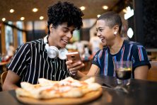 Two people look at a phone while they are at a restaurant with pizza in the foreground.