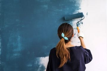 A woman paints a wall in her home blue. She has her hair tied up in a blue ribbon and is wearing a navy blue shirt.
