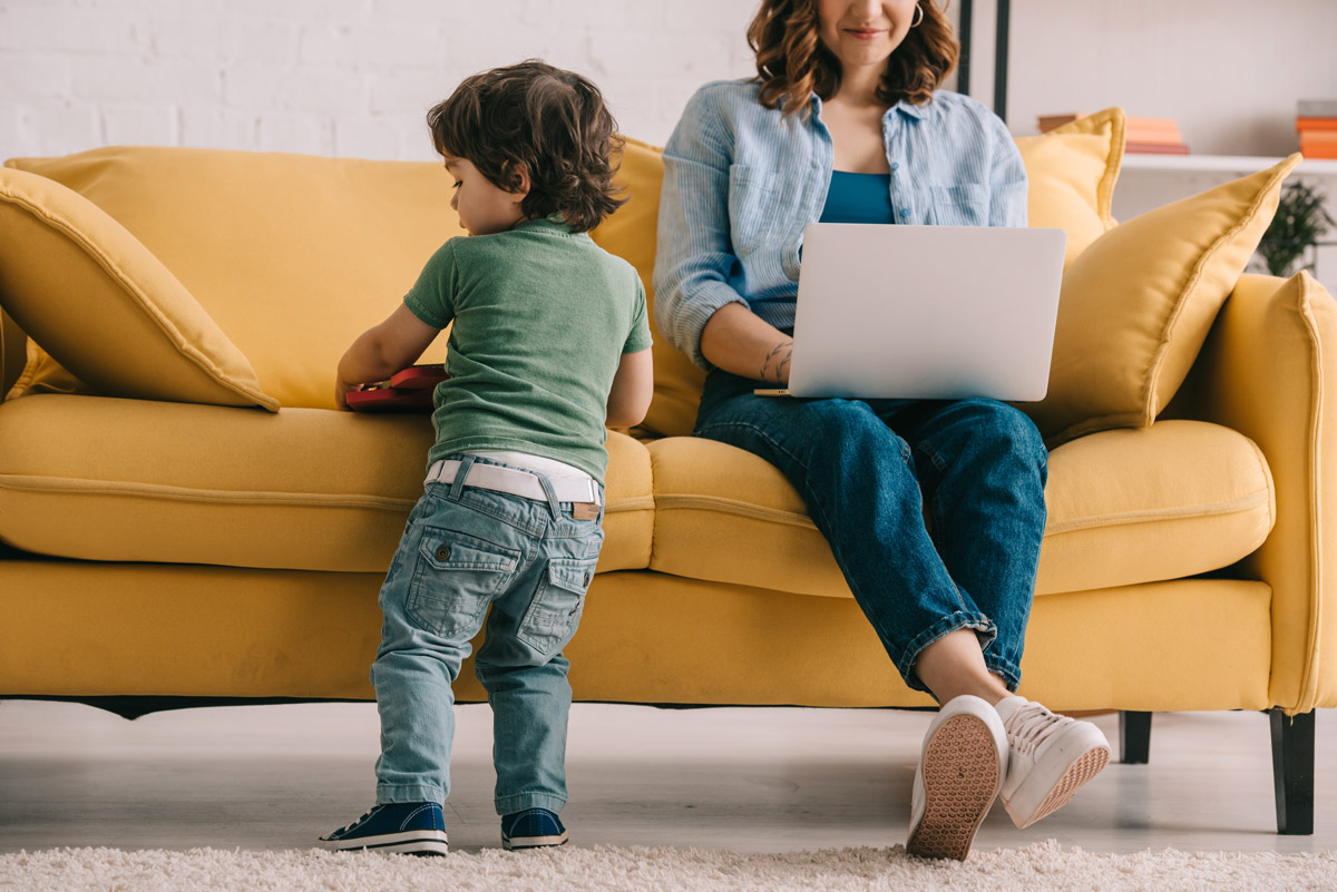 A woman uses her laptop while sitting on a yellow couch. Her son plays with a toy beside her.