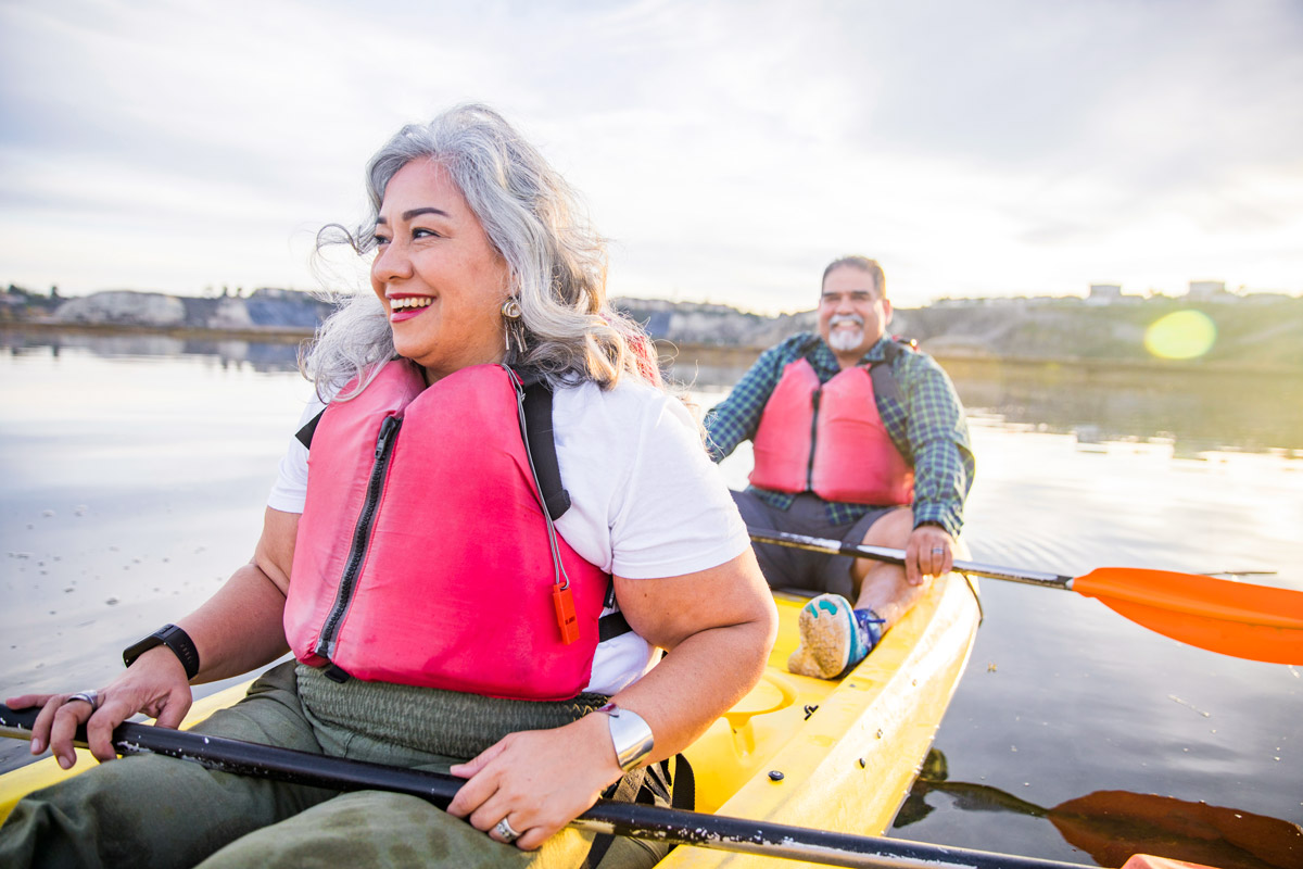 Two senior citizens kayak in a river wearing life vests.