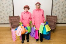 Two senior citizens wear pink coats and a pink hat as they hold multiple shopping bags in their hands.