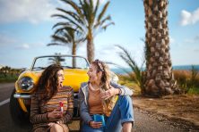 Two women lean against a yellow car while eating popsicles.